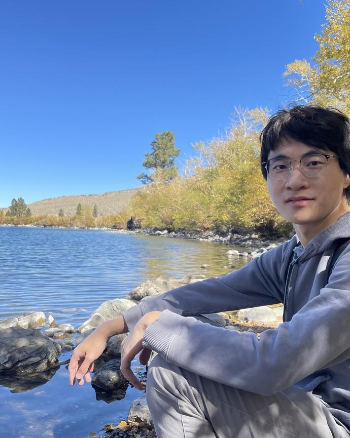 a man wearing glasses and a backpack poses on a rock in front of a body of water